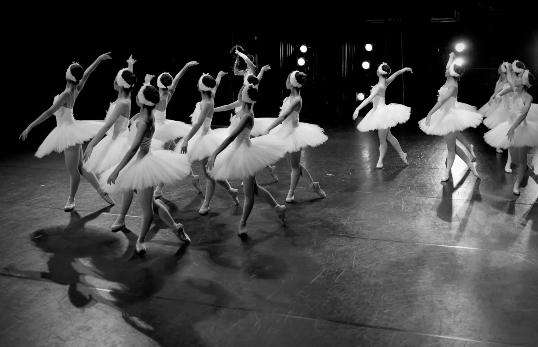 Zwart-wit foto van een groep balletdanseressen in tutu's op een donker podium, gevangen in een dynamische choreografie.